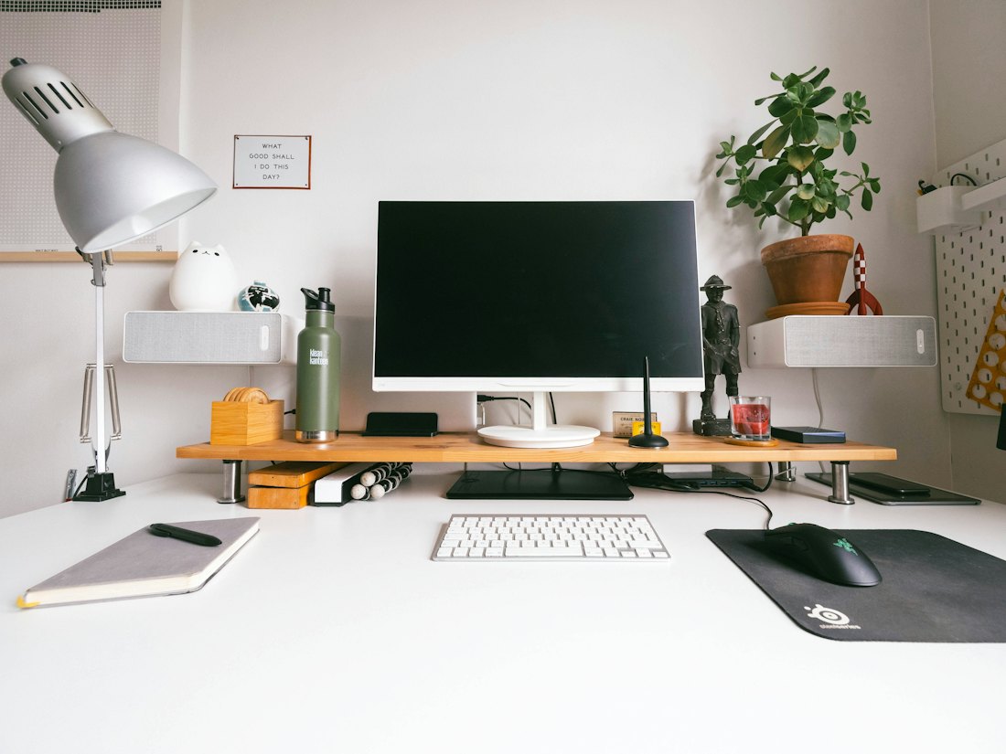 A bright desk with a monitor, keyboard, lamp and plants.